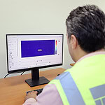 Çalışan, bilgisayar başında tehlike uyarı yeleğiyle bakanlık logosuyla, ofis ortamında teknik analiz yapıyor.