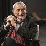 İki yaşlı adam ofis ortamında oturuyor, biri yüzünde gülümsemeyle sohbet ediyor, modern çalışma alanı.