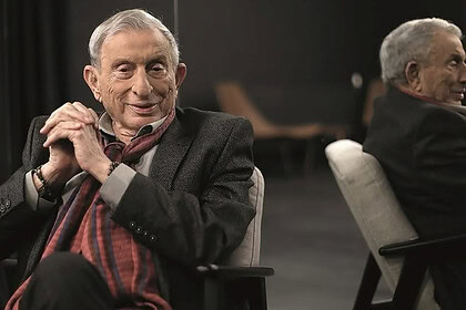 İki yaşlı adam ofis ortamında oturuyor, biri yüzünde gülümsemeyle sohbet ediyor, modern çalışma alanı.