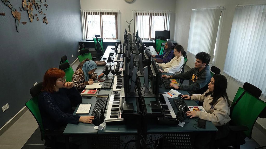 Alt text: Yeni nesil müzisyenler ve öğretmenler, modern kayıt stüdyosunda gitar ve klavye ile çalışıyor.