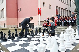 Başkan Yasin Özlü, şampiyon kız takımıyla satranç oynarken, büyük ilgiyle izleyen kalabalık ve çocuk.